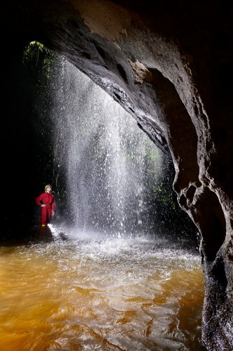 Caverna das Maos (Para, Brésil) - Cascade au soleil près de l'entrée de la grotte avec spéléo en fond et vasque d'eau jaune  en premier plan (verticale)(SP-23-1251 )